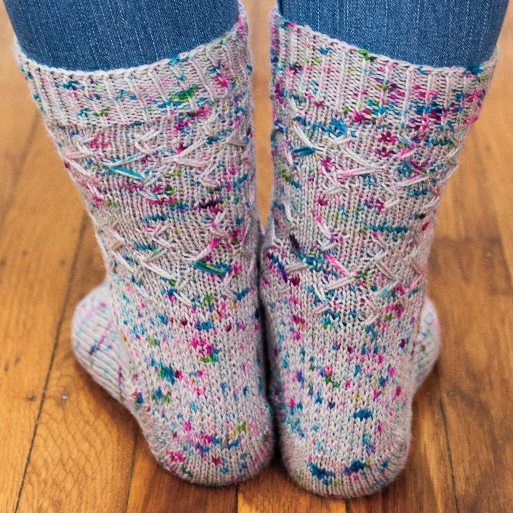 Back view of feet wearing grey knitted socks with blue and purple speckles and an angular slipped-stitch surface texture.