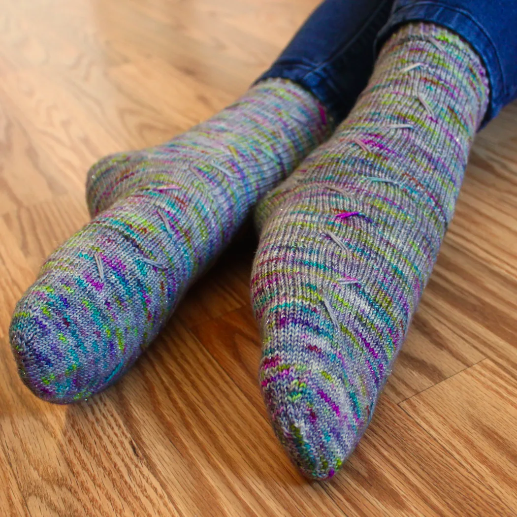 Crossed feet wearing grey socks with rainbow speckles that have lines of diagonal elongated stitches in two columns.