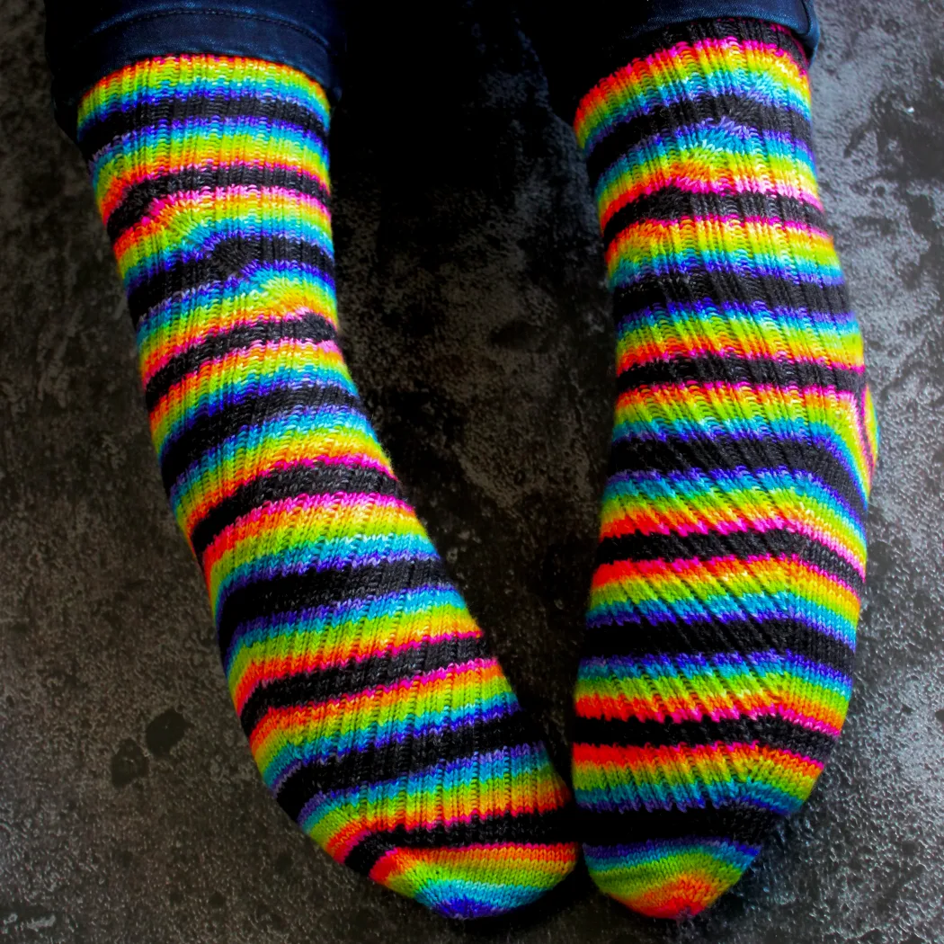 View from above of feet wearing black and rainbow striped socks with mirrored diagonal ribbing on the foot that twists to vertical on the leg.