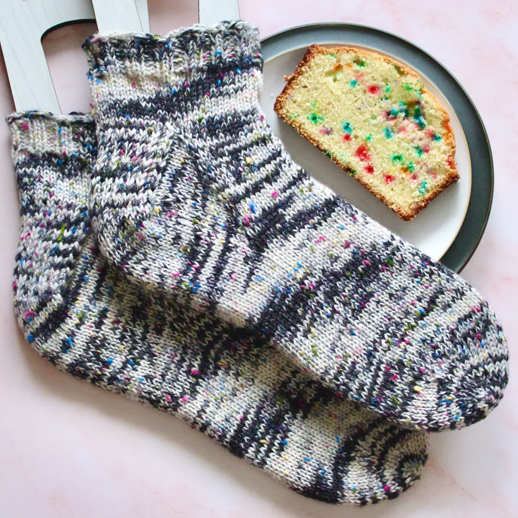 Wide view of black and white heavyweight socks with rainbow flecks on sock blockers next to a plate holding a slice of rainbow-speckled pound cake.