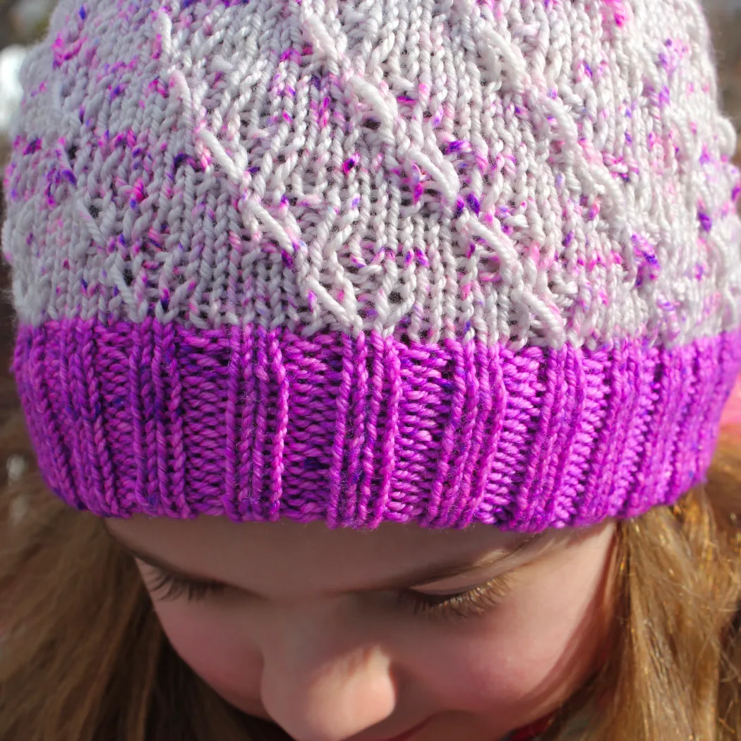 Close-up front view of child wearing white and neon purple toque with spiralling elongated-stitch texture and thick ribbed cuff.