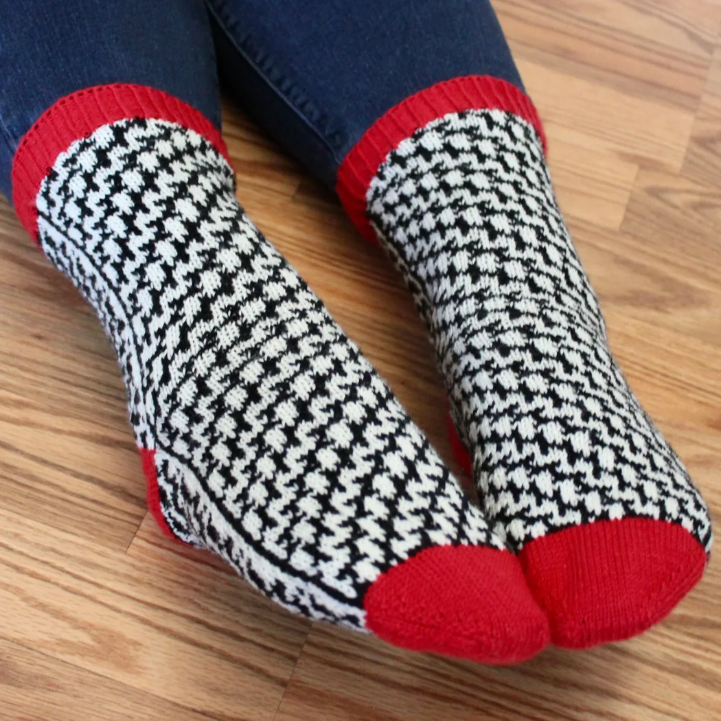 Feet wearing stranded colourwork socks with black and white mirrored optical-illusion lines and red heels, toes, and cuffs.