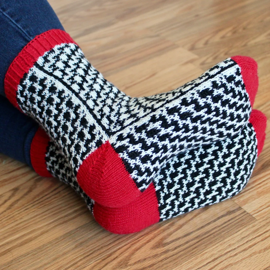 View of soles of stranded colourwork socks with black and white mirrored optical-illusion lines and red heels, toes, and cuffs.