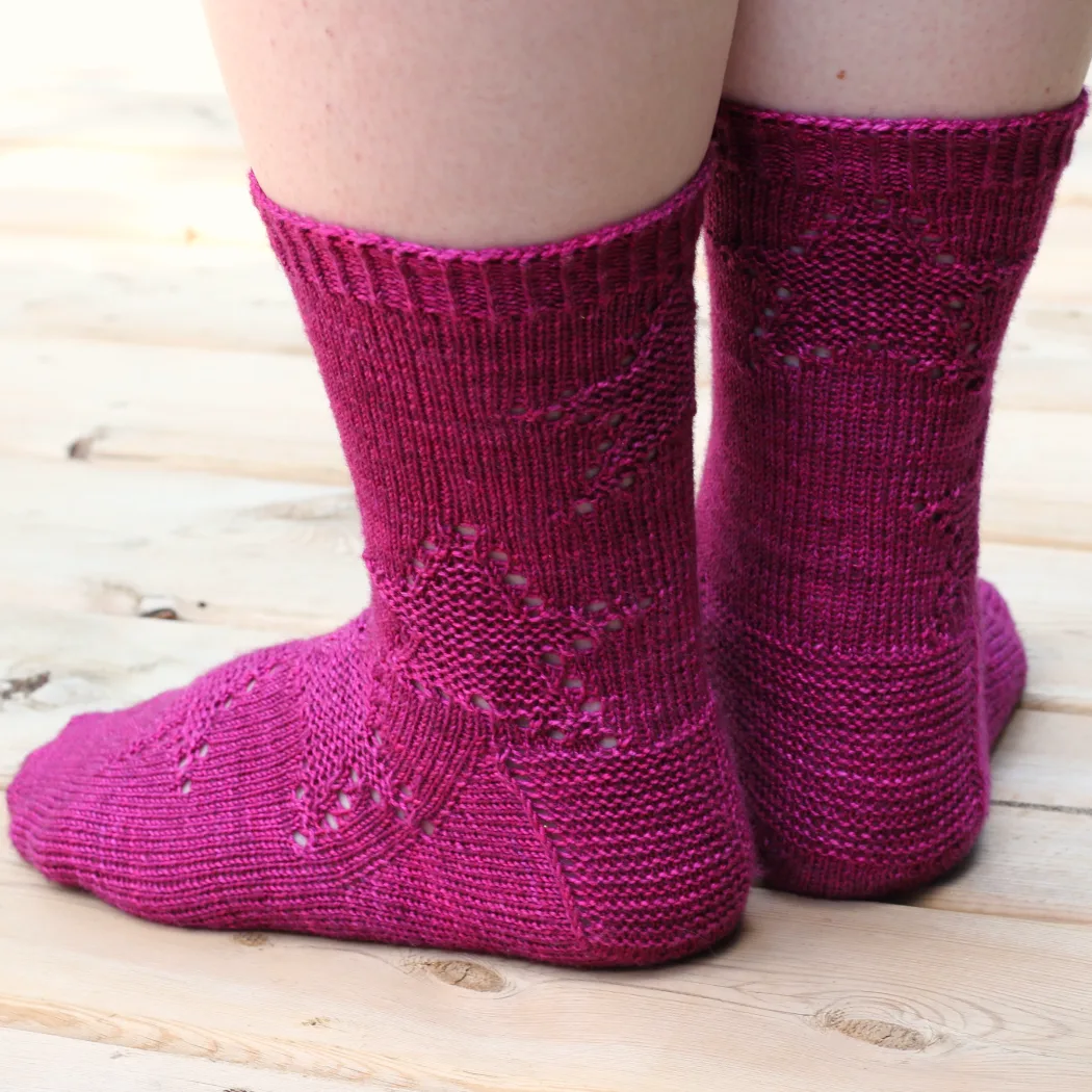 Back view of feet wearing dark pink knitted socks with spiralling star motif in texture and eyelets.