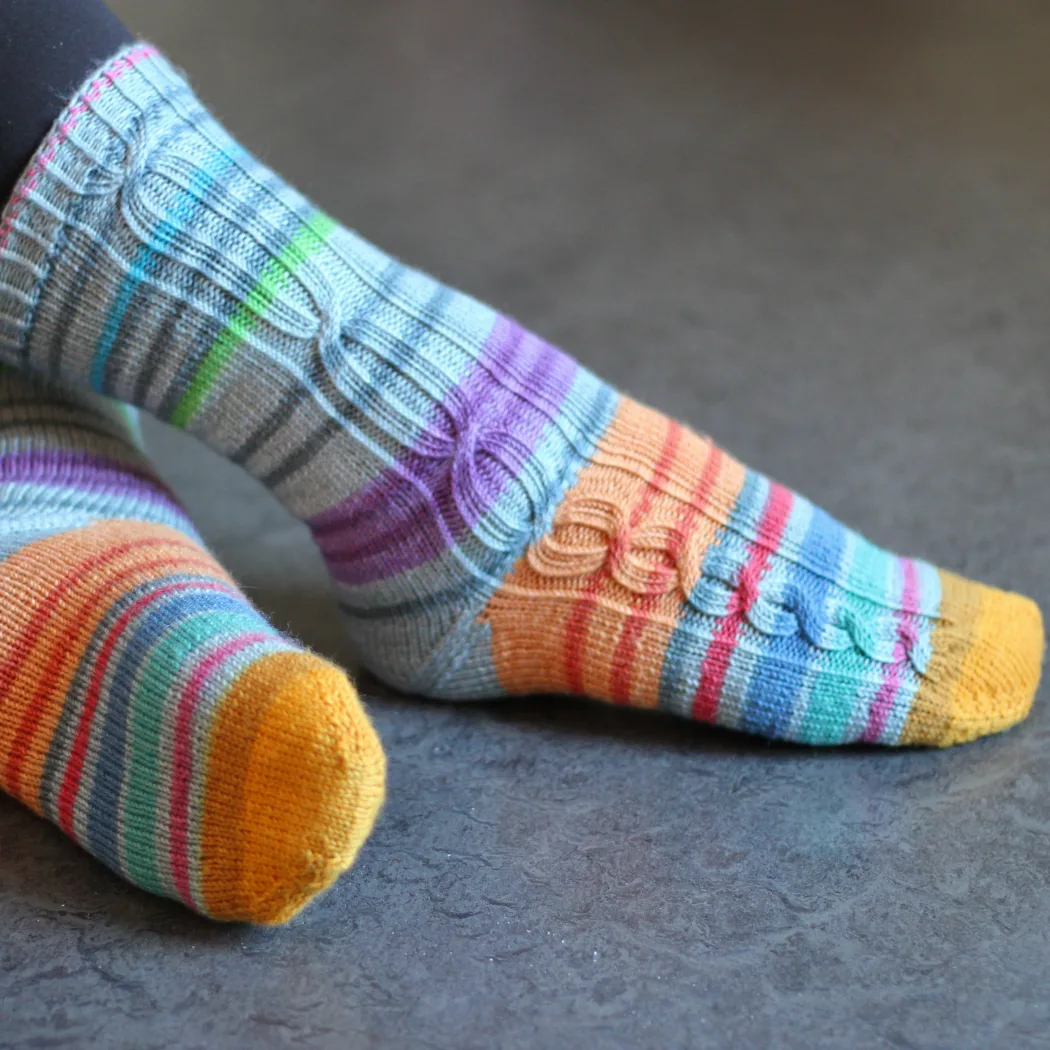 Side view of feet wearing striped knitted socks that have gradually-lengthening cables in panels on the outsides of the socks.