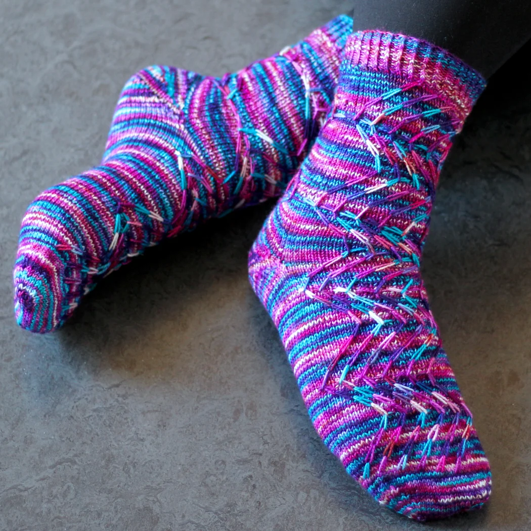 Crossed feet wearing pink, blue, purple, and white knitted socks with prominent zigzag slipped-stitch cable texture and short-row heel.