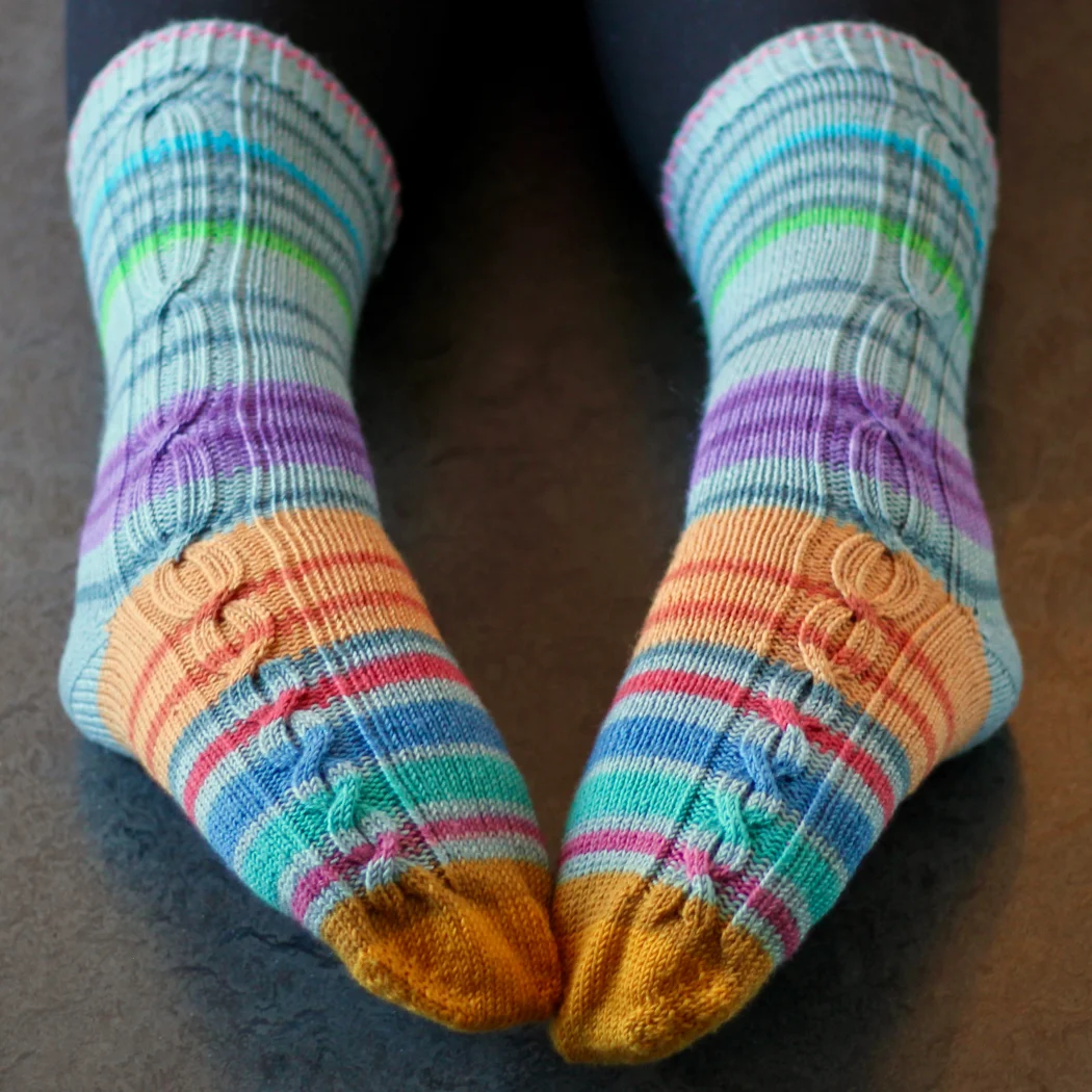 Feet wearing striped knitted socks that have gradually-lengthening cables in panels on the outsides of the socks.