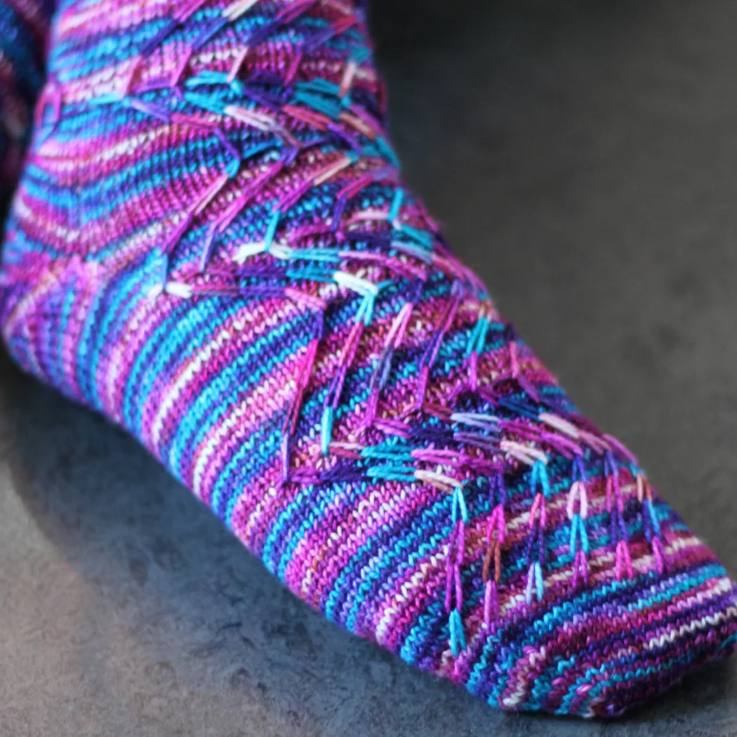 Close up of pink, blue, purple, and white knitted sock with prominent zigzag slipped-stitch cable texture.