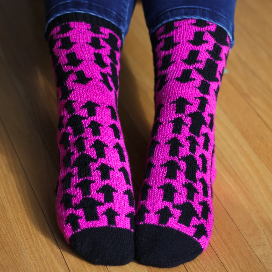 Feet wearing black and pink stranded colourwork socks with mirrored arrow motifs pointing down on the leg and up on the foot.