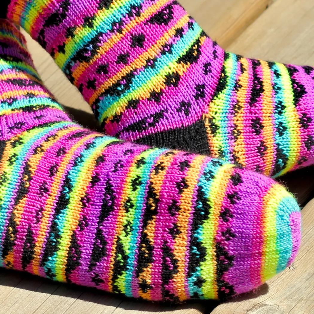 Feet wearing stranded colourwork socks with black heels and 8-bit sunglasses and sparkles motif on neon rainbow striped background.