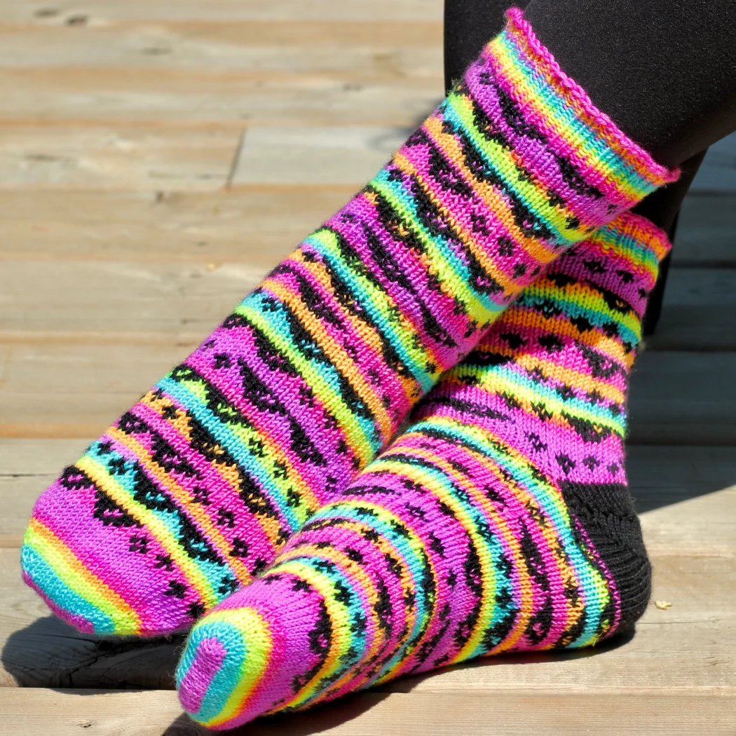 Feet wearing stranded colourwork socks with black heels and 8-bit sunglasses and sparkles motif on neon rainbow striped background.