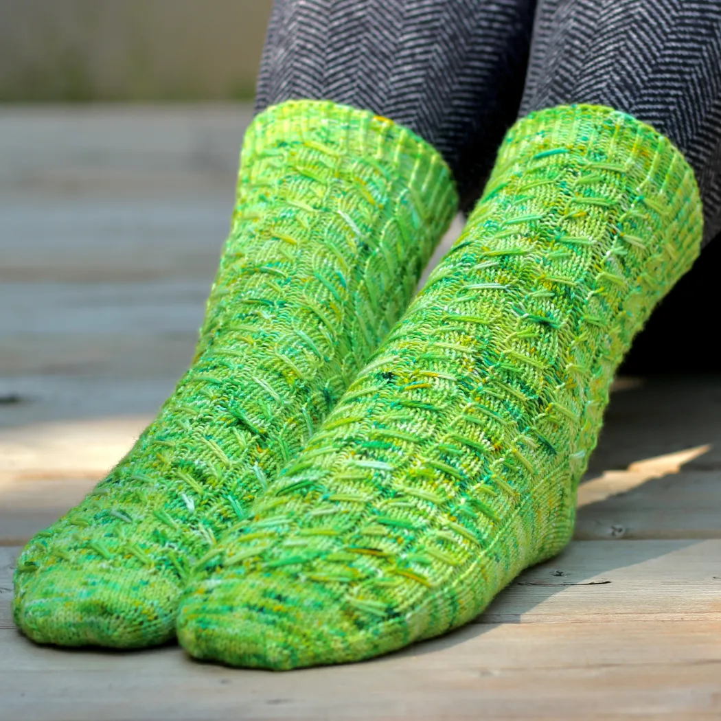 Side-by-side view of feet wearing green knitted socks with mirrored variable-width elongated-stitch cables in wide ribbing.