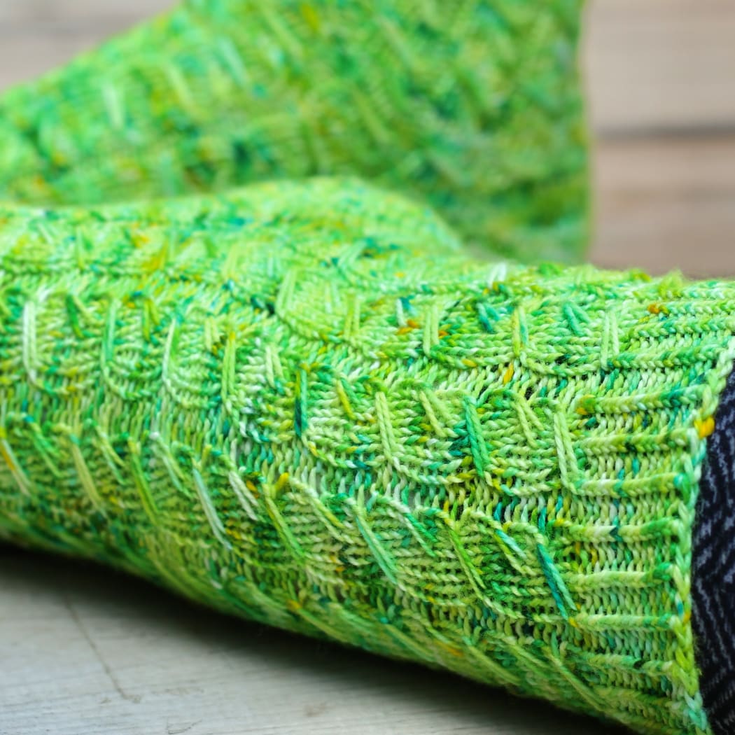 Close up of green knitted socks with variable-width elongated-stitch cables in wide ribbing that flow into the cuff ribbing.