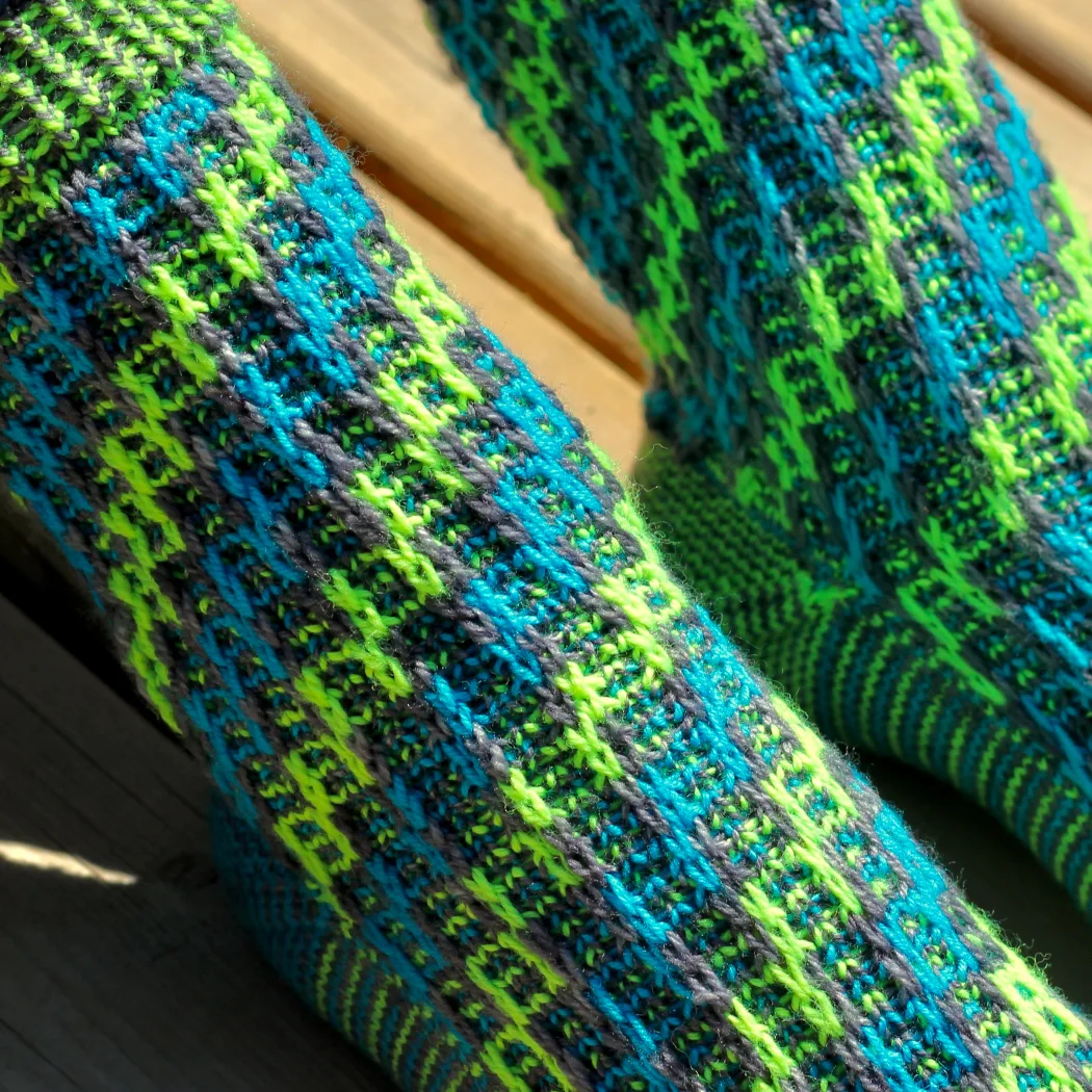 Feet wearing grey, blue, and neon green knitted socks with spiralling ladder motif.