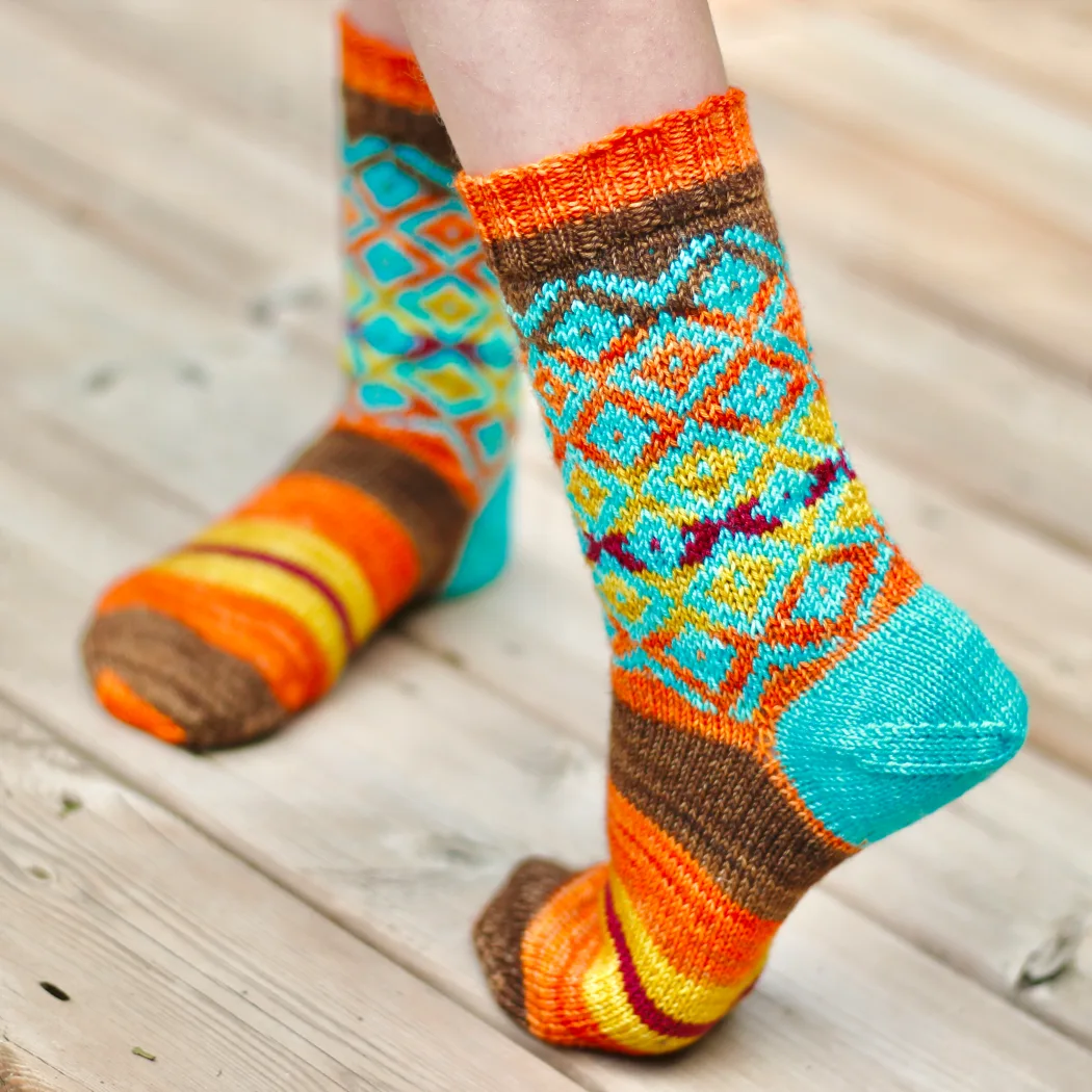 Twisting feet wearing brown, yellow, orange, and red striped knitted sock with overlaid bright blue diamonds motif on leg and bright blue heel.