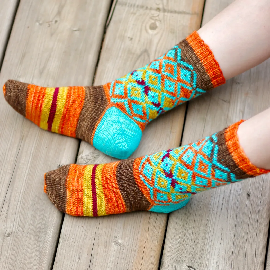 Top and side view of feet wearing brown, yellow, orange, and red striped knitted sock with overlaid bright blue diamonds motif on leg and bright blue heel.