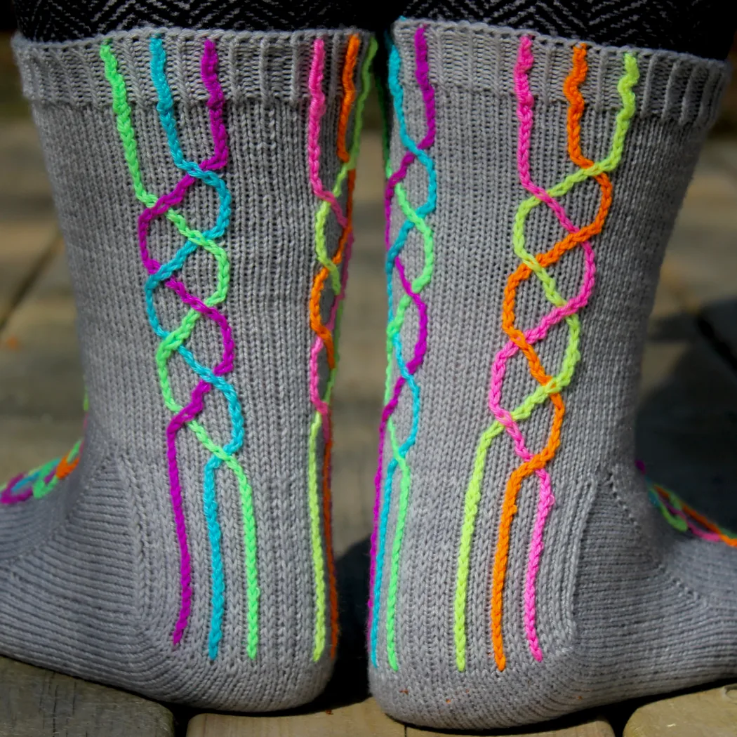 Back view of feet wearing grey knitted socks with two neon rainbow braid motifs.