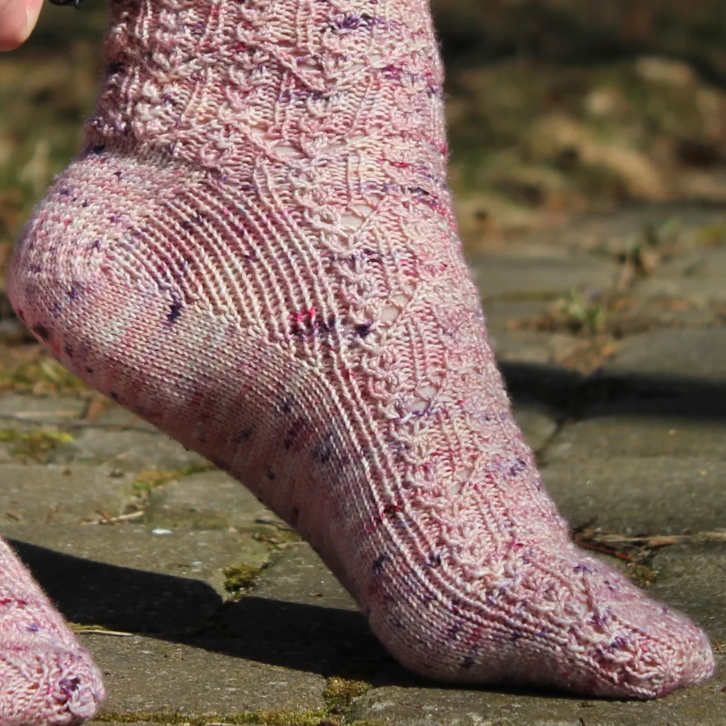 Foot on demi-pointe wearing pink knitted sock with delicate lace motif with spiralling stitches and ribbed gussets.