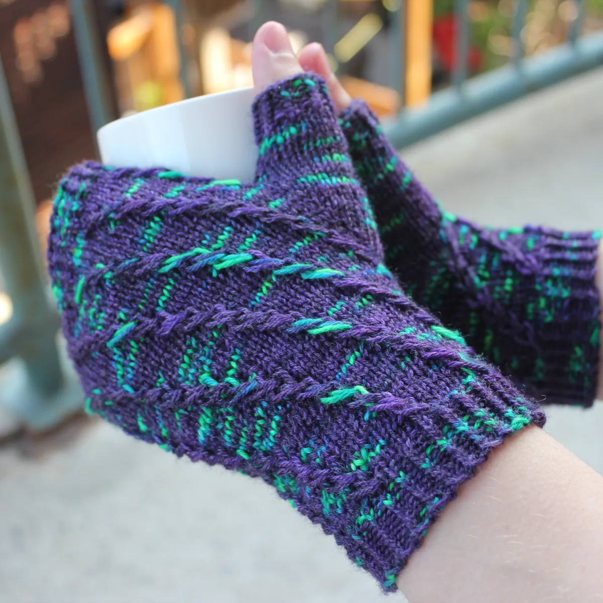 Side view of hands holding a white mug and wearing dark purple fingerless mitts with bright green flecks in a twisting spiralling pattern.