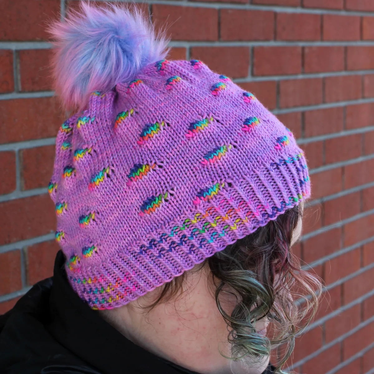 Woman wearing bright purple hat with pompom and randomly-distributed rainbow sections.