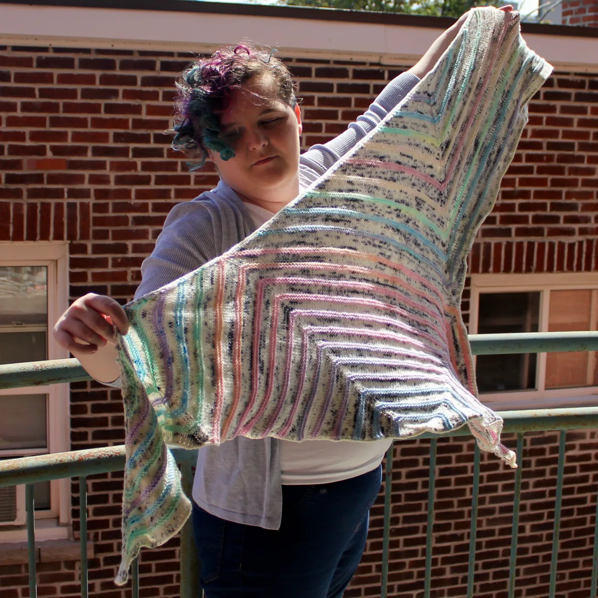 Woman holding a triangular shawl with flat white and black speckled sections between bumpy pastel stripes as it floats in the wind.