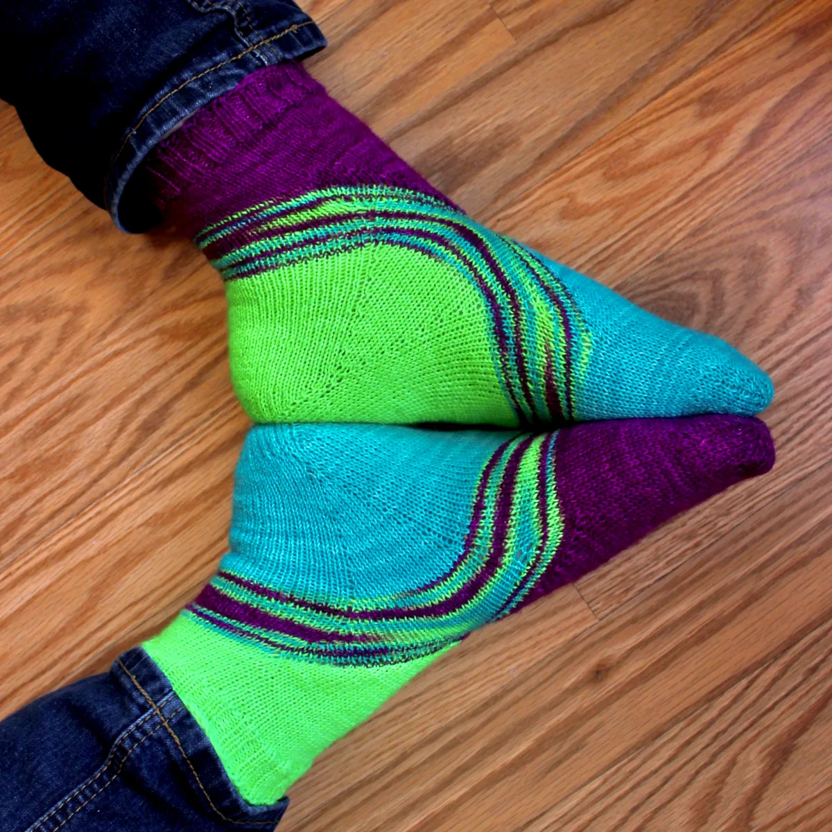 Feet lying sole-to-sole wearing bright green, blue, and purple socks with a multicoloured stripe between the colours.