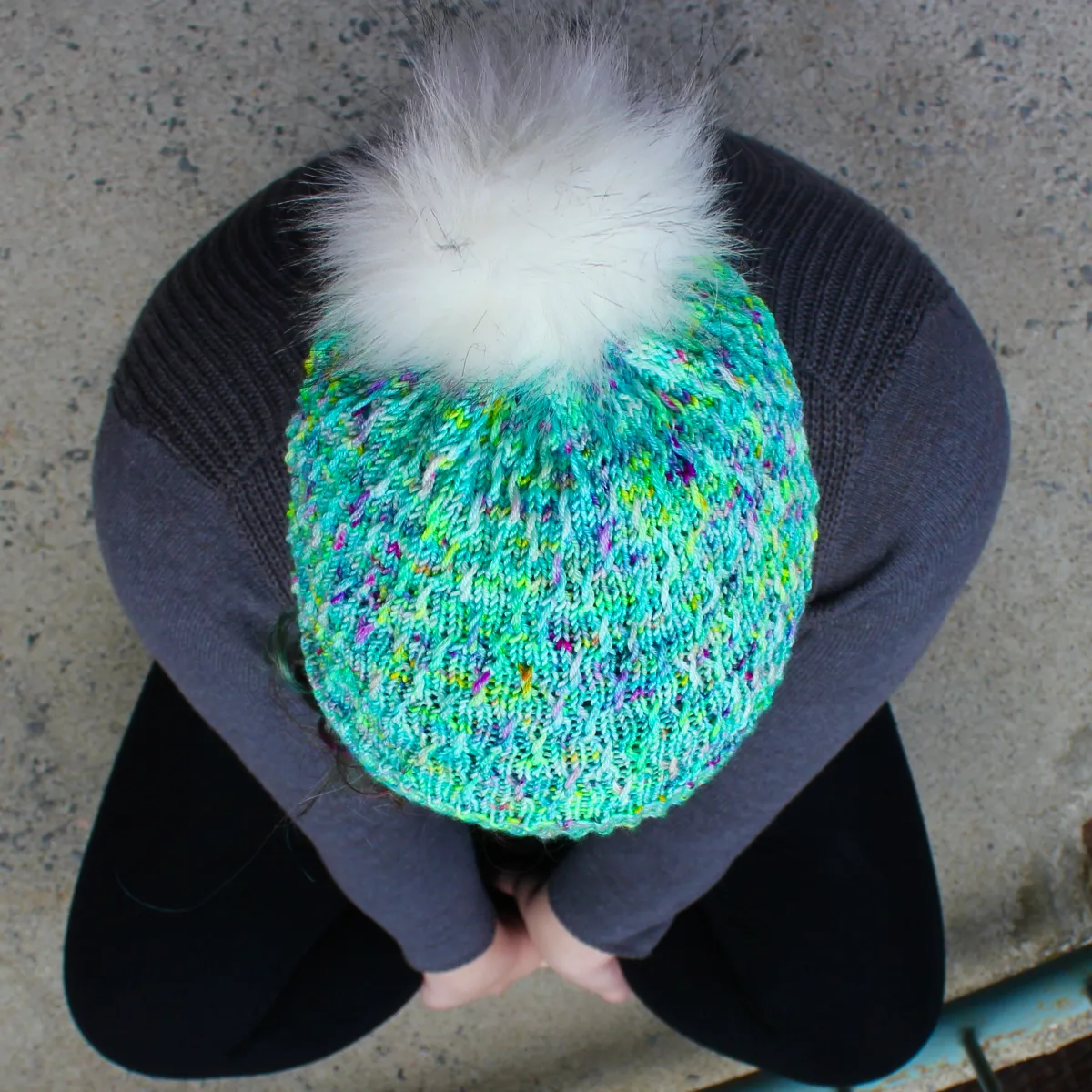 Top-down view of woman wearing bright green and rainbow hat with white pompom and twisted surface texture.