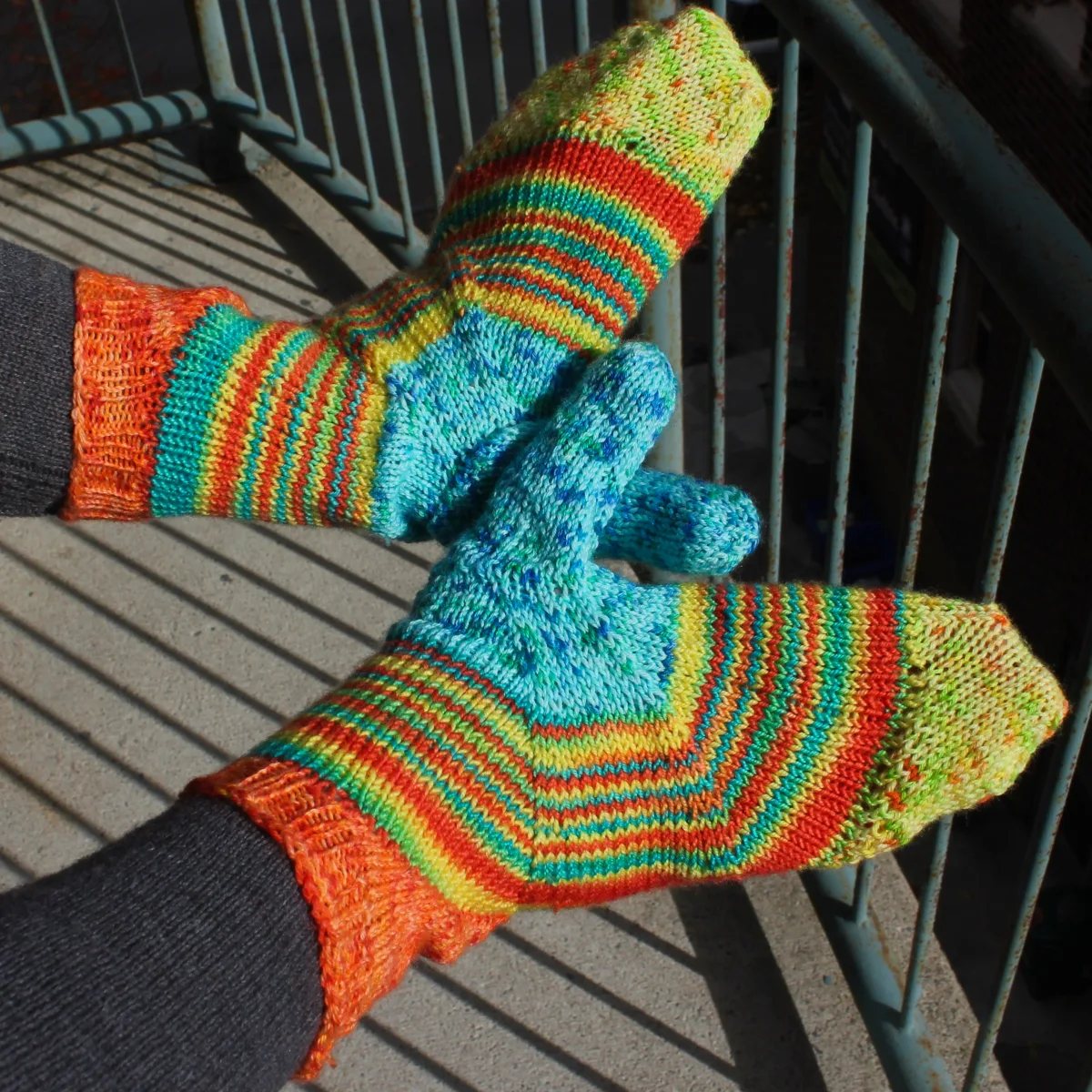 Top-down view of backs of hands wearing bright yellow, blue, and orange mittens with a multicoloured stripe across the palm between the colours.