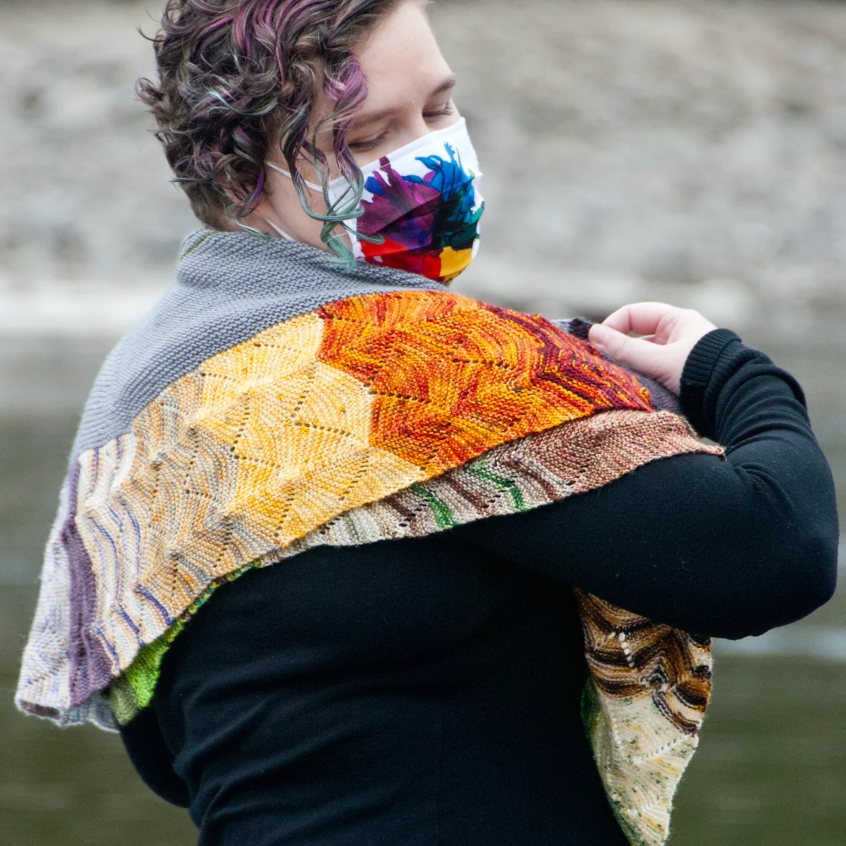 Woman in a mask wearing a large, colourful crescent-shaped knitted shawl with undulating textured lines.