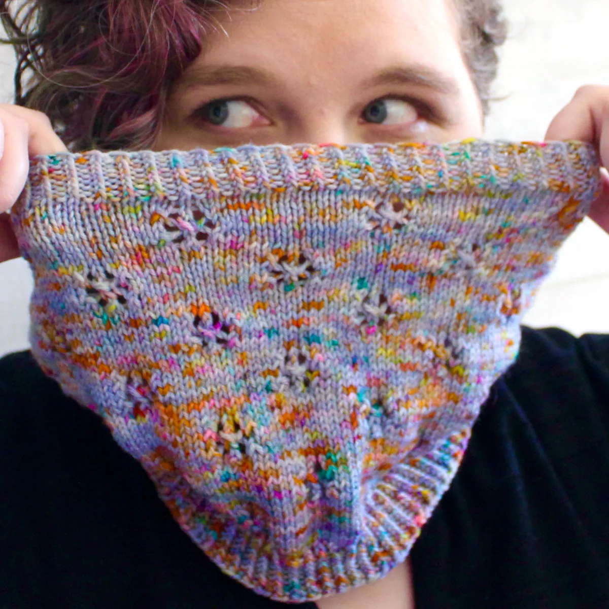 Woman holding up cowl that is light purple and rainbow-speckled knitted fabric with lace detail in front of her face.