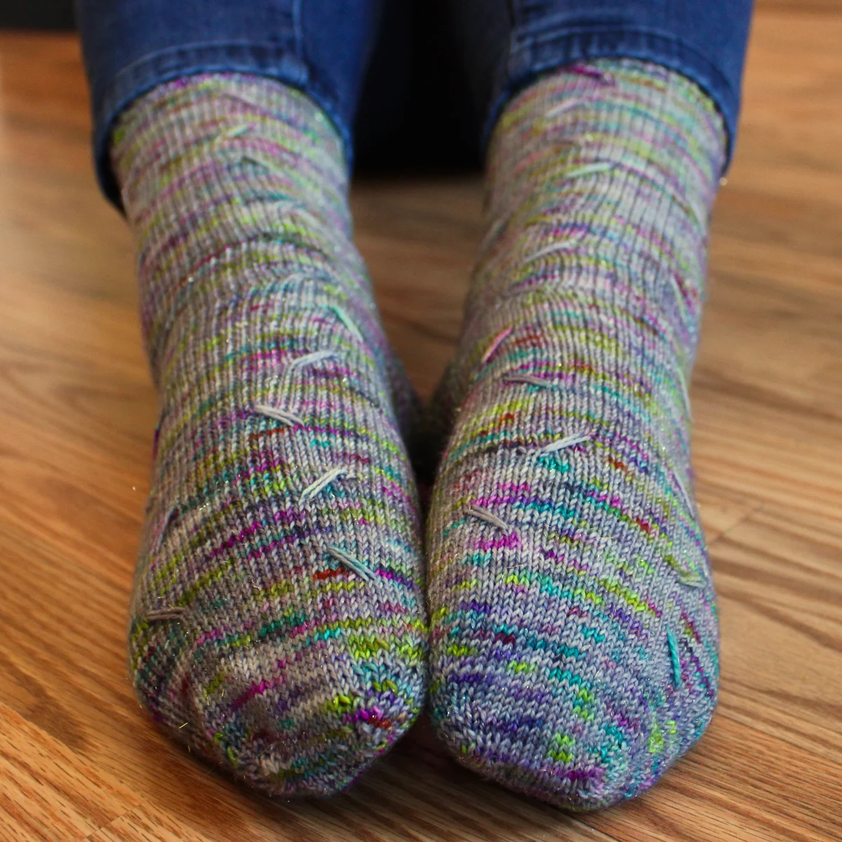 Toe up view of feet wearing grey socks with rainbow speckles that have lines of diagonal elongated stitches in two columns.