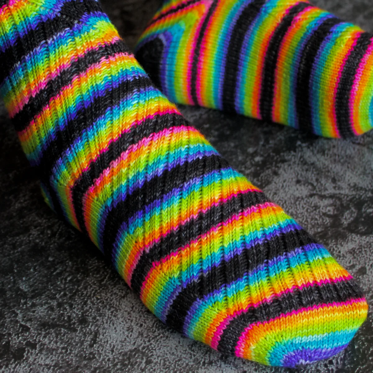 Close side view of feet wearing black and rainbow striped socks with diagonal ribbing on the foot that twists to vertical on the leg.