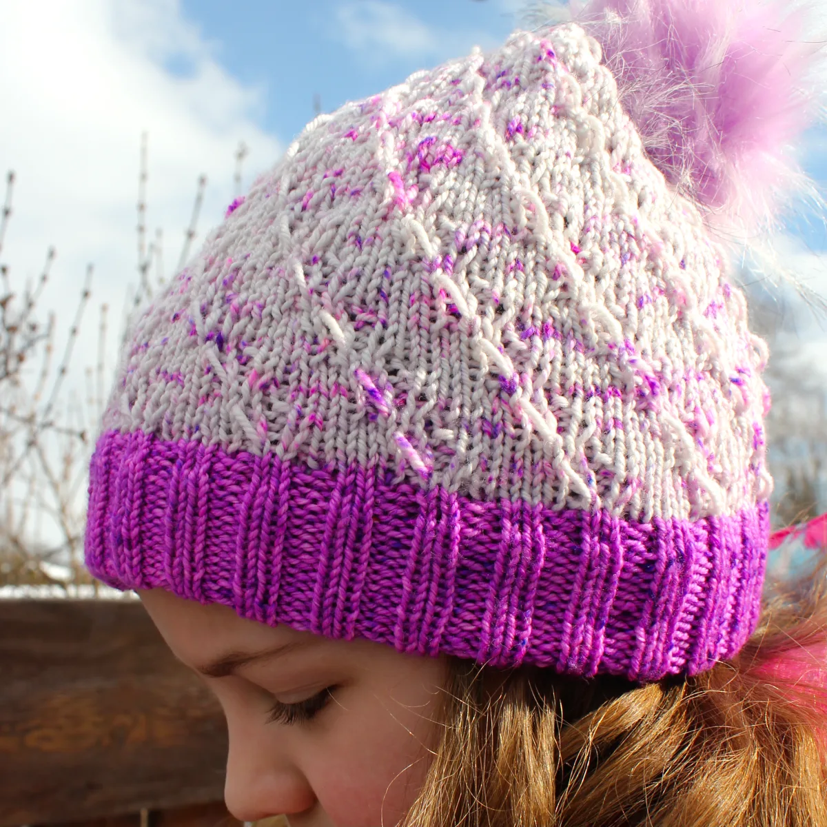 Side view of child wearing white and neon purple toque with spiralling elongated-stitch texture and light purple pompom.