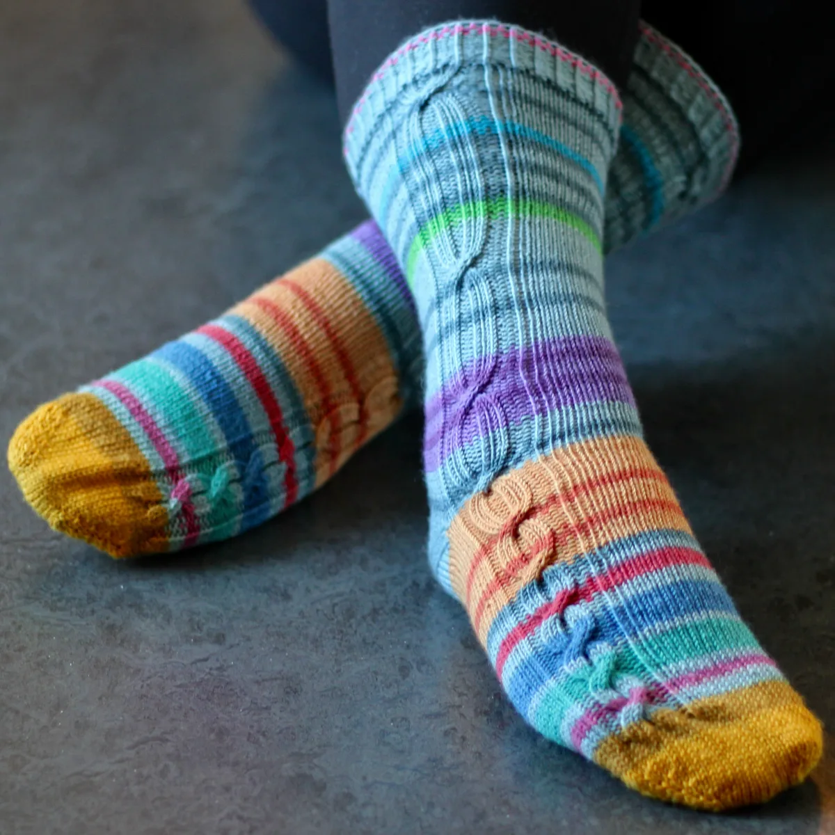 Crossed feet wearing striped knitted socks that have gradually-lengthening cables in panels on the outsides of the socks.