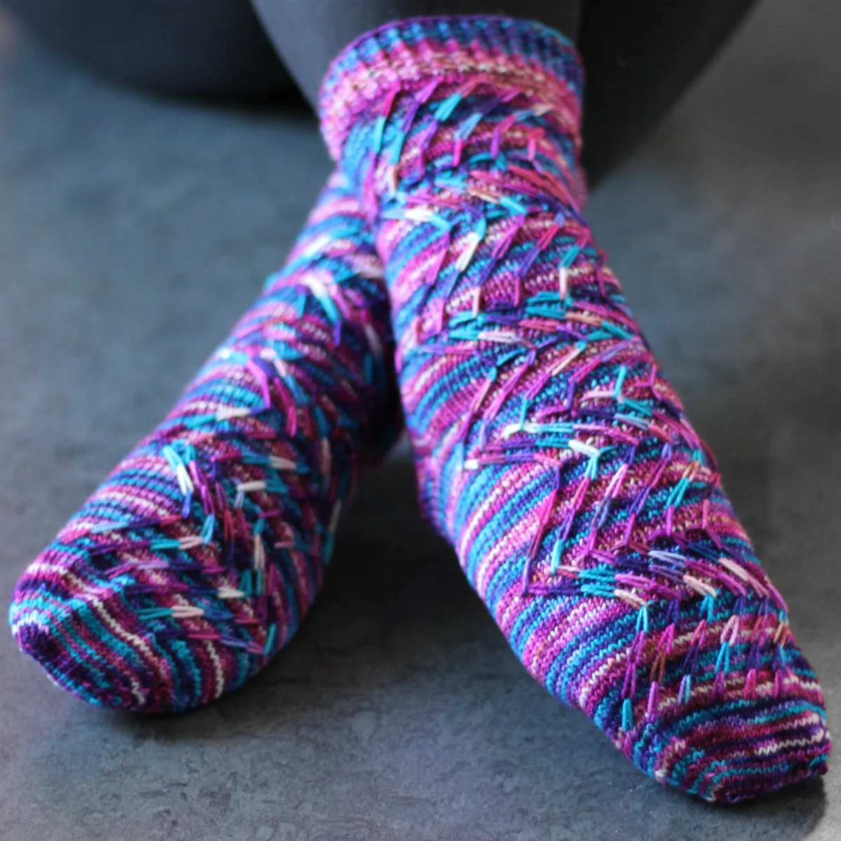 Crossed feet wearing pink, blue, purple, and white knitted socks with prominent zigzag slipped-stitch cable texture.