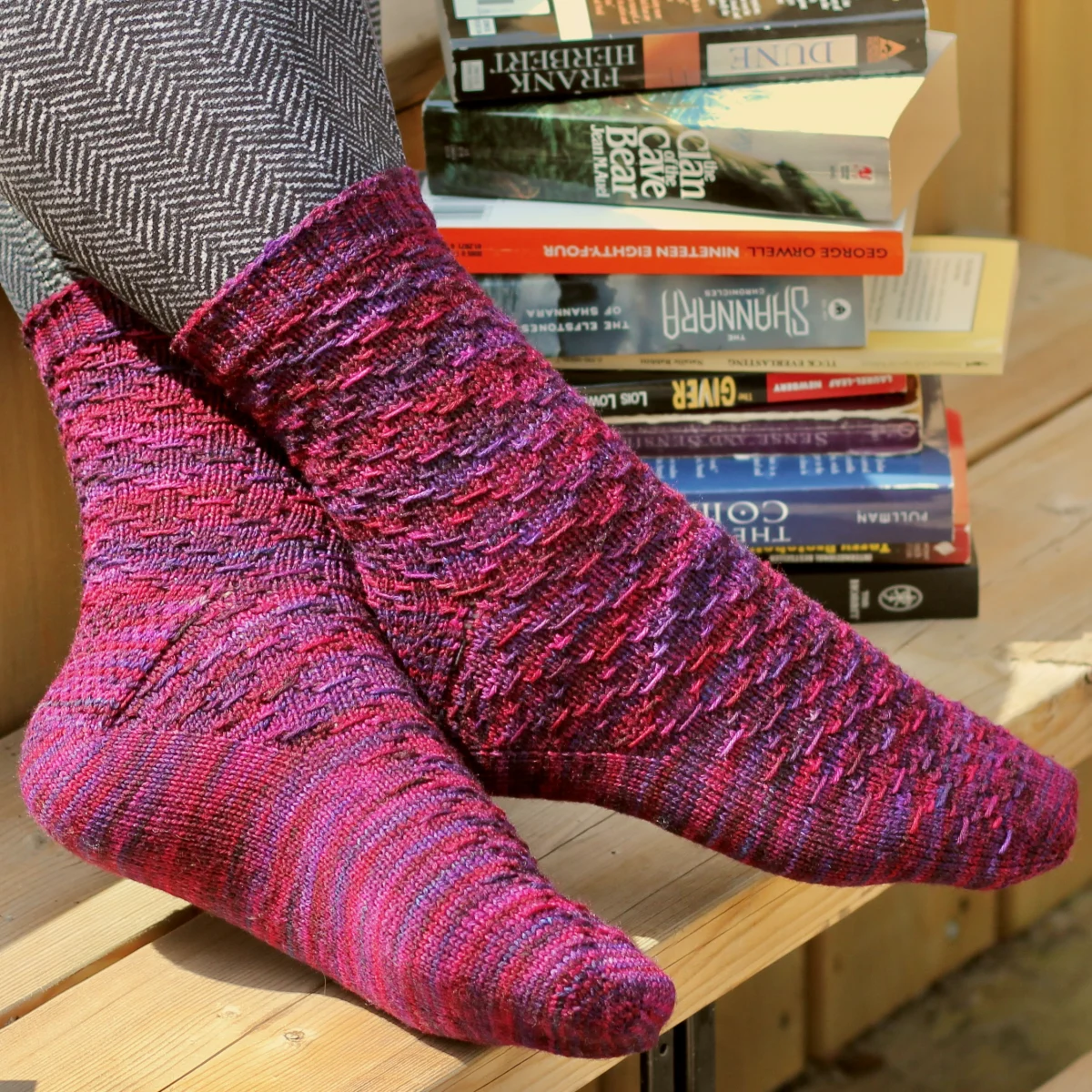 Crossed feet wearing red and purple socks with horizontal diamond-shaped surface texture with novels in the background.