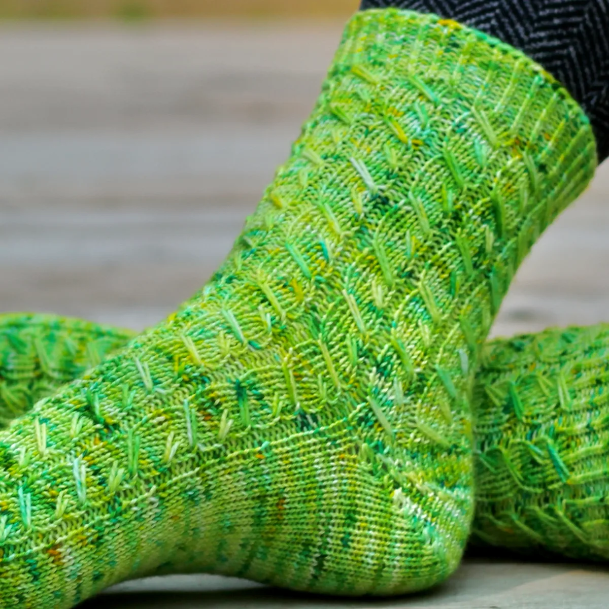 Crossed feet with front foot turned to the side wearing green knitted socks with variable-width elongated-stitch cables in wide ribbing.