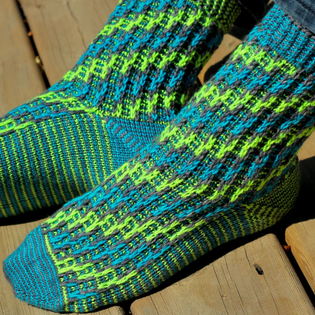 Feet wearing grey, blue, and neon green knitted socks with spiralling ladder motif.