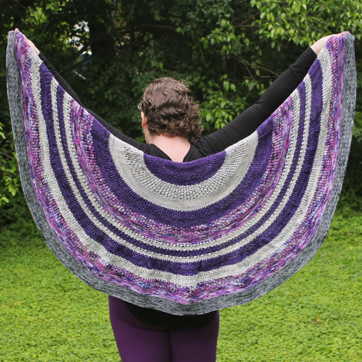 Woman holding up a purple and grey knitted shawl with lace sections, irregular stripes, and a textured edge.