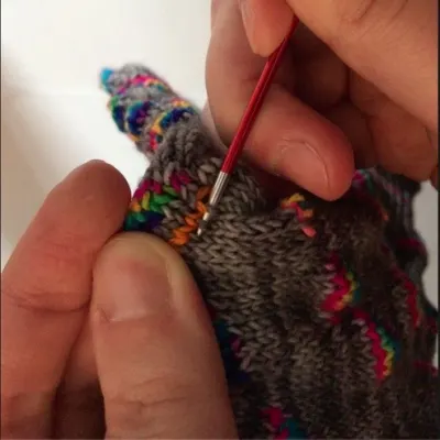 Hands holding knitted fabric and using a double-pointed needle to pick up stitches from it.