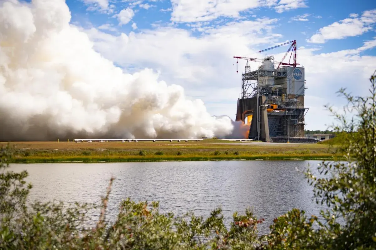 A massive cloud of white vapor billows out from a structure where an RS-25 engine is firing. The flames are just barely visible through the structure. In the foreground is a body of water.