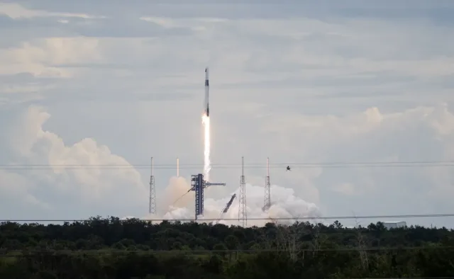 A SpaceX Falcon 9 rocket carrying the company's Dragon spacecraft launches. There is a bright column of fire under the rocket as it moves upward. Clouds of vapor spread out below the rocket, almost blending into the similarly shaped clouds in the sky.