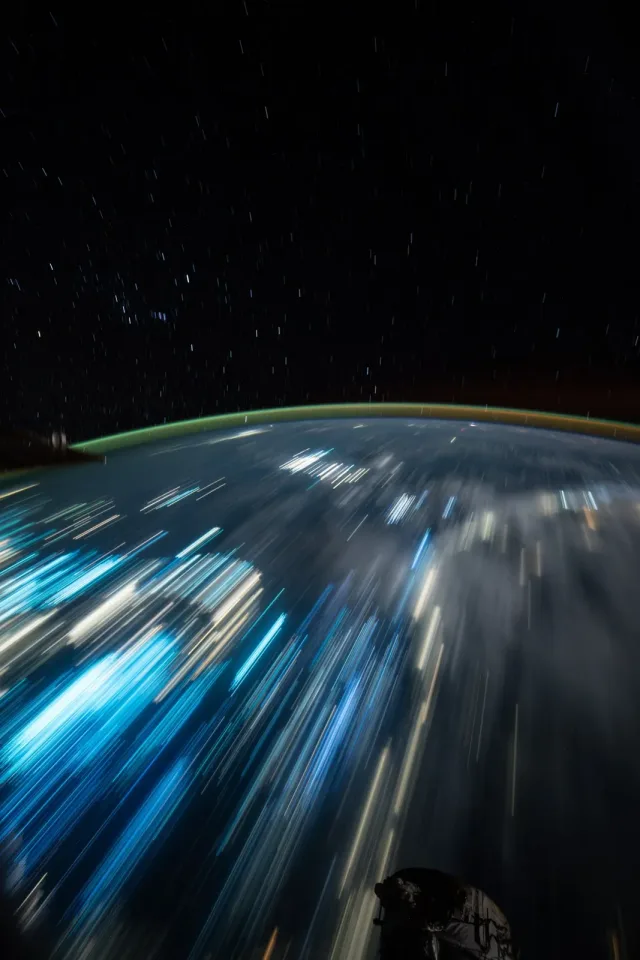 Streaks of blue and yellow lights cover Earth. Above, faint streaks of light made by stars can be seen.