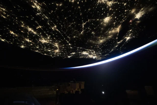A dark Earth, lit up in areas by city lights, takes up most of the image. A small sliver of light is visible at the bottom right curve of our home planet. Part of the International Space Station can be faintly seen at the bottom of the photo.