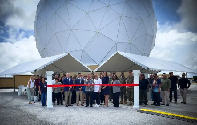 A large group gathers behind a red ribbon for a ribbon-cutting ceremony beneath white tents, with a large white geodesic radome in the background.
