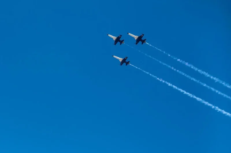 Looking up, three white and black fighter jets fly in formation across a bright, blue sky.