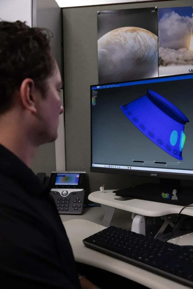 A person sits at a workstation viewing a computer monitor that displays a blue 3D wireframe engineering model of a spacecraft component.
