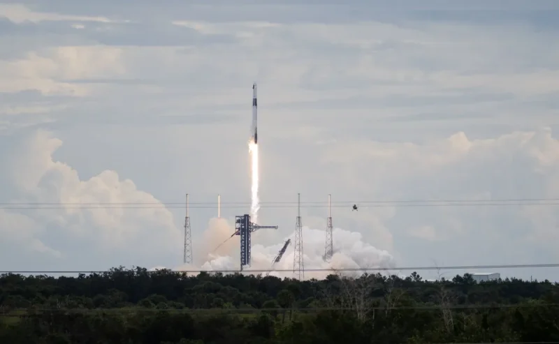A SpaceX Falcon 9 rocket carrying the company's Dragon spacecraft launches. There is a bright column of fire under the rocket as it moves upward. Clouds of vapor spread out below the rocket, almost blending into the similarly shaped clouds in the sky.