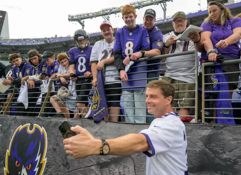 Reid Wiseman holds up a black phone to take a selfie with Baltimore Ravens fans at a football game. The fans are dressed in Ravens jerseys and shirts in purple and white. The fans are in the stands above, while Wiseman is on the field just below.