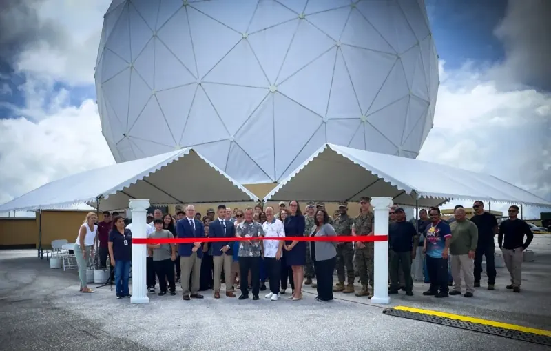 A large group gathers behind a red ribbon for a ribbon-cutting ceremony beneath white tents, with a large white geodesic radome in the background.